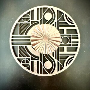 May include: A circular object featuring a light-colored, laser-cut geometric pattern with straight lines and circles. Centered on top is a smaller, metallic copper-toned circular disc with a radial, sunburst-like texture of fine, embossed lines.