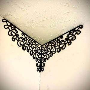 Puede incluir: Decoración de pared de esquina de metal negro con un diseño intrincado y simétrico. La pieza decorativa presenta un patrón repetitivo de formas arremolinadas, creando un aspecto estilizado y elegante. La decoración está montada en una pared y un techo blancos.