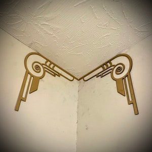 May include: Two decorative wooden corner moldings in a light brown color with a white swirl design. The moldings are installed in the corner of a room where the wall meets the ceiling.