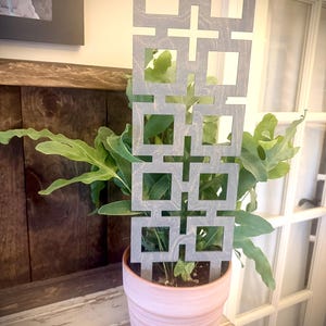 May include: A grey wooden trellis with a geometric square design is placed in a pink ceramic pot with a green plant. The trellis is in front of a wooden background and a white door.