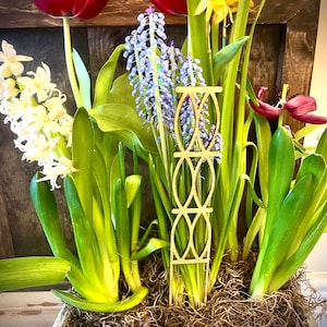 May include: A wooden plant stake with a decorative design is placed in a planter with a variety of flowers, including red tulips, white hyacinths, and blue grape hyacinths.