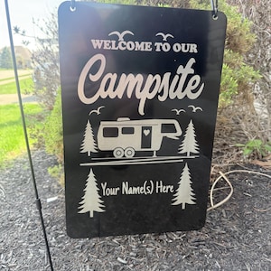 Peut inclure: Panneau en métal noir avec du texte blanc qui dit "Bienvenue à notre camping" avec un camping-car stylisé et des arbres. Le texte "Votre nom(s) ici" est sous le camping-car.