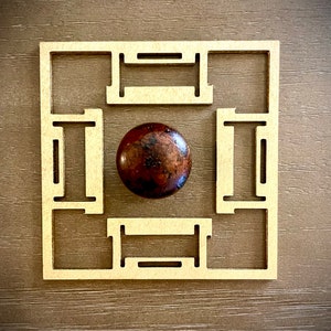 May include: A wooden square frame with a brown sphere in the center. The frame has a geometric design with four squares and four rectangles.