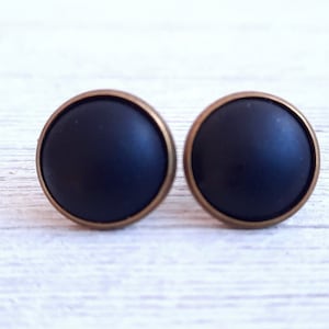 Könnte beinhalten: Ein Paar schwarze Ohrstecker mit goldfarbenem Rand. Die Ohrringe sind rund und haben eine matte Oberfläche.