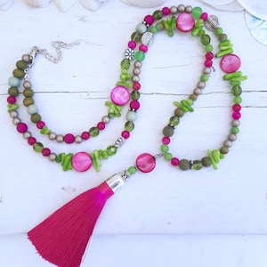 Può includere: Una lunga collana a più strati con perline rosa e verdi, accenti argentati e un fiocco rosa. La collana è realizzata con diversi materiali, tra cui vetro, metallo e legno.