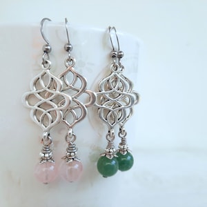 Op de afbeelding: Een paar zilveren oorhangers met een geometrisch ontwerp en twee edelsteen druppels. Eén oorhanger heeft twee roze edelsteen druppels en de andere heeft twee groene edelsteen druppels.