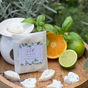 Può includere: Un set di sei cere per profumare l'ambiente di colore bianco a forma di foglie con fiori viola incorporati nella cera. Le cere sono su un vassoio di legno con una borsa bianca sullo sfondo. La borsa ha un'etichetta che dice "E&R Evie & Ruby Candle Co. Lime, Basil & Mandarin".