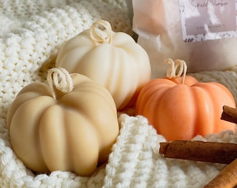 May include: Three pumpkin-shaped candles in cream, beige, and orange colors, each featuring a coiled wick at the top. A white paper bag with a label reading Spiced Pumpkin is visible, along with two brown cinnamon sticks in the foreground.