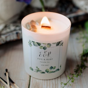 Könnte beinhalten: Eine weiße Kerze mit einem Holzdocht und einem Etikett mit der Aufschrift "E&R Evie & Ruby Candle Co. SPA DAY". Die Kerze ist von Grünpflanzen und Streichhölzern umgeben.