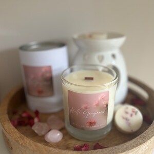 Pode incluir: Uma bandeja de madeira com uma vela branca em um pote de vidro, um aquecedor de cera branco de cerâmica e duas velas de cera brancas. O pote da vela tem um rótulo rosa com o texto "Hello Gorgeous" e as palavras "The Love & Ruby Candle Co.". A bandeja também tem pétalas de rosa rosa, dois cristais de quartzo rosa e uma vela branca em uma lata.