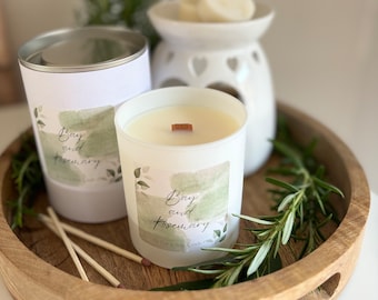 May include: Two candles with a wooden wick, one in a glass jar and one in a metal tin, both with a white label that says "Bay and Rosemary" in a handwritten font. The candles are on a wooden tray with sprigs of rosemary.