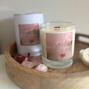 Puede incluir: Un tarro de vidrio para velas con una etiqueta blanca que dice "Hello Gorgeous" y "By Evie & Ruby Candle Co.". La vela está sobre una bandeja de madera con pétalos de rosa rosa y cristales de cuarzo rosa.