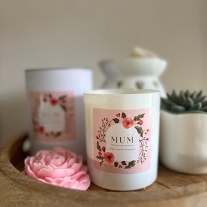 May include: Two white candles with floral labels. The label on the front candle says "MUM" with the text "Thank you for being you." The label on the back candle is blurry. The candles are on a wooden tray with a pink rose-shaped soap.