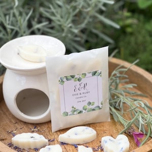 Könnte beinhalten: Ein Set mit sechs weißen Wachs-Schmelzen in Blatt- und Herzformen mit getrockneten Lavendelblüten. Die Wachsschmelzen sind in einem weißen Papierbeutel mit einem Etikett verpackt, auf dem "E&R Evie & Ruby Candle Co. SPA DAY" steht.