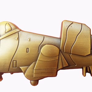 May include: A gold-colored metal pin shaped like an A-10 Warthog aircraft. The pin has a detailed design and a shark mouth painted on the front.