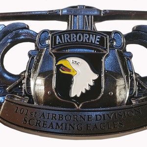 May include: A metal emblem featuring the words "AIRBORNE" and "101st AIRBORNE DIVISION SCREAMING EAGLES." The emblem has a central shield with a white eagle head and yellow beak. The design includes a helicopter and decorative wings.