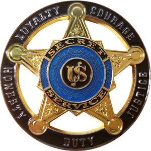 May include: A gold five-pointed star badge with a blue center circle. The circle has a black "US" with a dollar sign inside. The star has the words "SECRET SERVICE" around the center circle. The outer ring of the badge is black with the words "LOYALTY", "HONESTY", "COURAGE", "JUSTICE", and "DUTY" around the star.