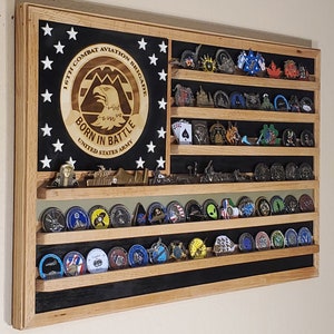 May include: A wooden shadow box with a black background and a white star pattern. The shadow box has a wooden frame and a glass front. The shadow box is decorated with a wooden plaque that says "167th Combat Aviation Brigade Born in Battle United States Army". The shadow box also has several shelves that are filled with military challenge coins.