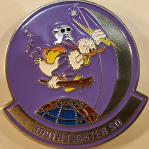 May include: A purple and silver metal coin with a cartoon duck holding a lightning bolt. The duck is wearing a purple robe and has a white bird on its back. The coin has the text "309th Fighter SQ" around the edge.