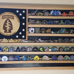 May include: A wooden shadow box with a black and white American flag background. The shadow box has five shelves with various military challenge coins displayed. The top shelf has a wooden plaque with the text "5TH SPECIAL FORCES GROUP AIRBORNE UNITED STATES ARMY".