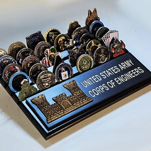 May include: A black wooden display case with a blue and silver inscription that reads "United States Army Corps of Engineers". The case is filled with a variety of military challenge coins.