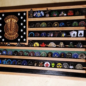 May include: Wooden display case shaped like a flag, featuring multiple shelves filled with collectible coins and pins. The left side has a black background with white stars and a military emblem. The case has a natural wood frame.