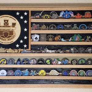May include: A wooden shadow box with a black background and multiple shelves. The shelves are filled with various challenge coins. The top shelf has a black and white American flag with the text "130th Engineer Brigade Combat Ready United States Army".