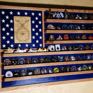 May include: A wooden shadow box with a blue velvet background and a US flag design. The shadow box has multiple shelves for displaying challenge coins. The top shelf has a gold plaque with the Department of the Air Force seal.