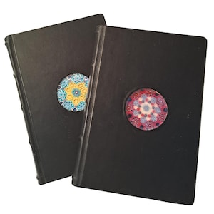 Può includere: Due diari neri con disegni circolari decorativi. Un diario presenta un design con elementi gialli, blu e bianchi, mentre l'altro ha un motivo rosso, rosa e blu. I diari sono rilegati con cuciture visibili.