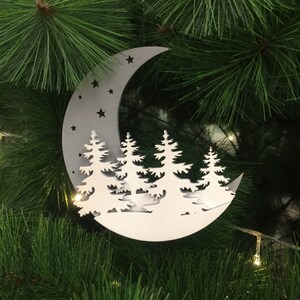 Puede incluir: Un adorno de madera blanco con forma de media luna con fondo gris. La luna tiene estrellas recortadas y una silueta de un bosque con cinco árboles.