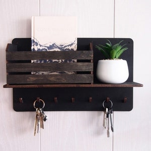 Puede incluir: Porta llaves negro con una repisa y una pequeña caja de madera. La caja contiene un libro titulado "catch the waves" y hay una pequeña planta en maceta en la repisa. Hay cinco ganchos en la parte inferior del porta llaves.