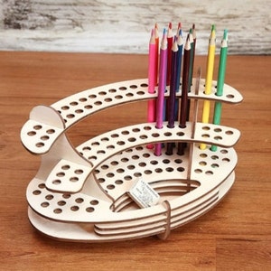 Könnte beinhalten: Ein Bleistifthalter aus Holz mit mehreren Ebenen und Löchern zum Aufbewahren von Bleistiften. Der Halter ist aus hellem Holz und hat eine abgerundete Form. Es befinden sich mehrere farbige Bleistifte im Halter.