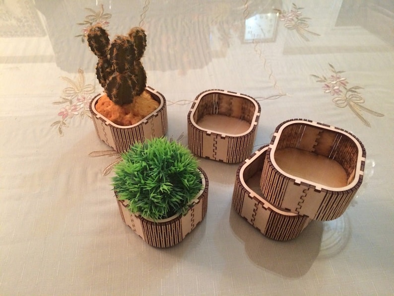 Puede incluir: Cuatro macetas de madera, dos apiladas una encima de la otra. Una maceta tiene una peque&ntilde;a planta de cactus dentro. Las macetas est&aacute;n hechas de finas tiras de madera que se unen para formar una forma cuadrada.