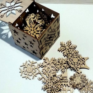 May include: A wooden box with a snowflake design on the lid, open to reveal a collection of laser-cut wooden snowflakes. The snowflakes are scattered on a white surface.