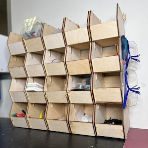 Può includere: Un organizzatore di stoccaggio a parete in legno con più scomparti. L'organizzatore è realizzato in legno chiaro e ha un design a nido d'ape. È riempito con vari piccoli oggetti, tra cui fili, strumenti e altri rifornimenti.