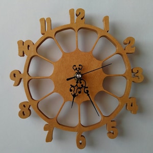 May include: A wooden clock with a unique design featuring 12 numbers around the edge and a decorative clock face. The clock hands are black and the clock face is a light brown color.