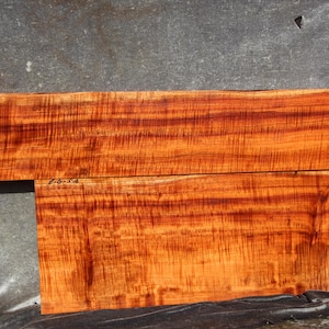 Puede incluir: Dos tablas de madera rectangulares con un tono rojo anaranjado vibrante y un patrón de veta ondulado prominente. La tabla superior presenta el texto escrito a mano "F-G-5-B" y la inferior muestra el texto "Z-G-5-A" en tinta negra.