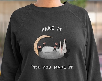 Könnte beinhalten: Schwarzes Sweatshirt mit dem Text "FAKE IT 'TIL YOU MAKE IT" und einer Cartoon-Opossum-Illustration. Das Opossum liegt auf dem Rücken. Das Sweatshirt hat eine dunkle Farbe.