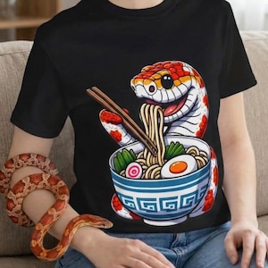 May include: Black t-shirt with a cartoon snake design eating ramen from a blue bowl. The snake is red, white, and orange. The bowl has a white and blue geometric pattern. The shirt is worn with blue jeans.