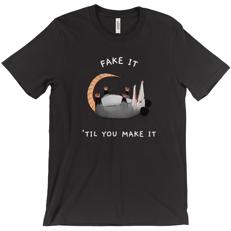 K&ouml;nnte beinhalten: Schwarzes T-Shirt mit einem Cartoon-Opossum, das sich totstellt, mit dem Text "FAKE IT" dar&uuml;ber und "'TIL YOU MAKE IT" darunter. Das Opossum ist grau mit einem beigen Schwanz und rosa Krallen. Ein humorvolles Grafik-T-Shirt.