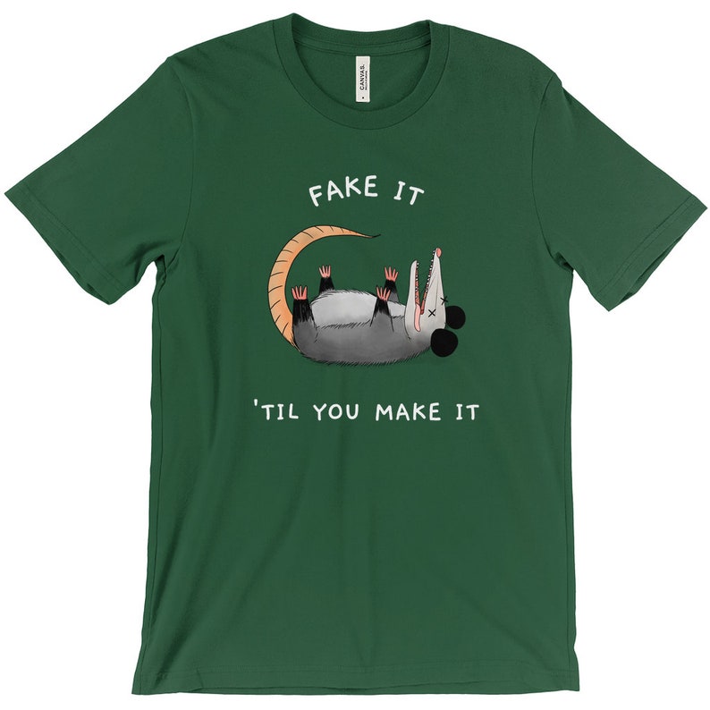 K&ouml;nnte beinhalten: Dunkelgr&uuml;nes T-Shirt mit dem Text "FAKE IT 'TIL YOU MAKE IT" und einem Cartoon-Opossum. Das Opossum ist grau mit einem beigen Schwanz und rosa F&uuml;&szlig;en. Das T-Shirt ist aus einem weichen Material.