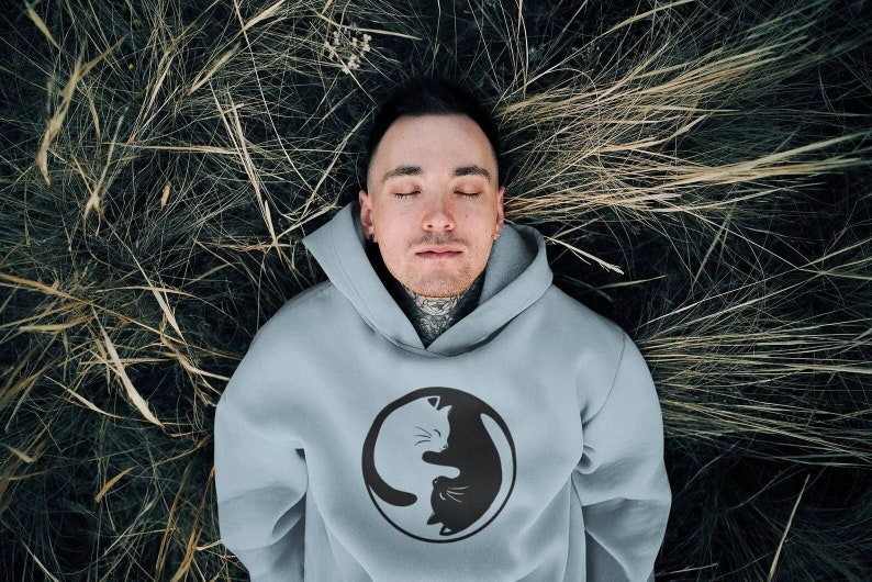 May include: A light blue hoodie featuring a black and white yin-yang cat design. The hoodie is made of a soft material and has a hood and a front pocket. The person in the image is lying in tall grass.