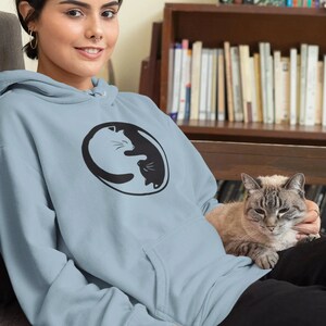 May include: A light blue hoodie featuring a black and white yin-yang cat design. The hoodie has a kangaroo pocket and a drawstring hood. The person in the image is wearing black pants and is sitting with a cat.
