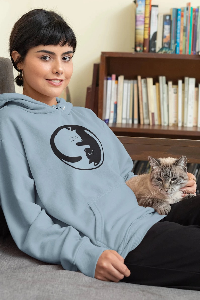 May include: A light blue hoodie featuring a black and white yin-yang cat design. The hoodie has a kangaroo pocket and a drawstring hood. The person in the image is wearing black pants and is sitting with a cat.