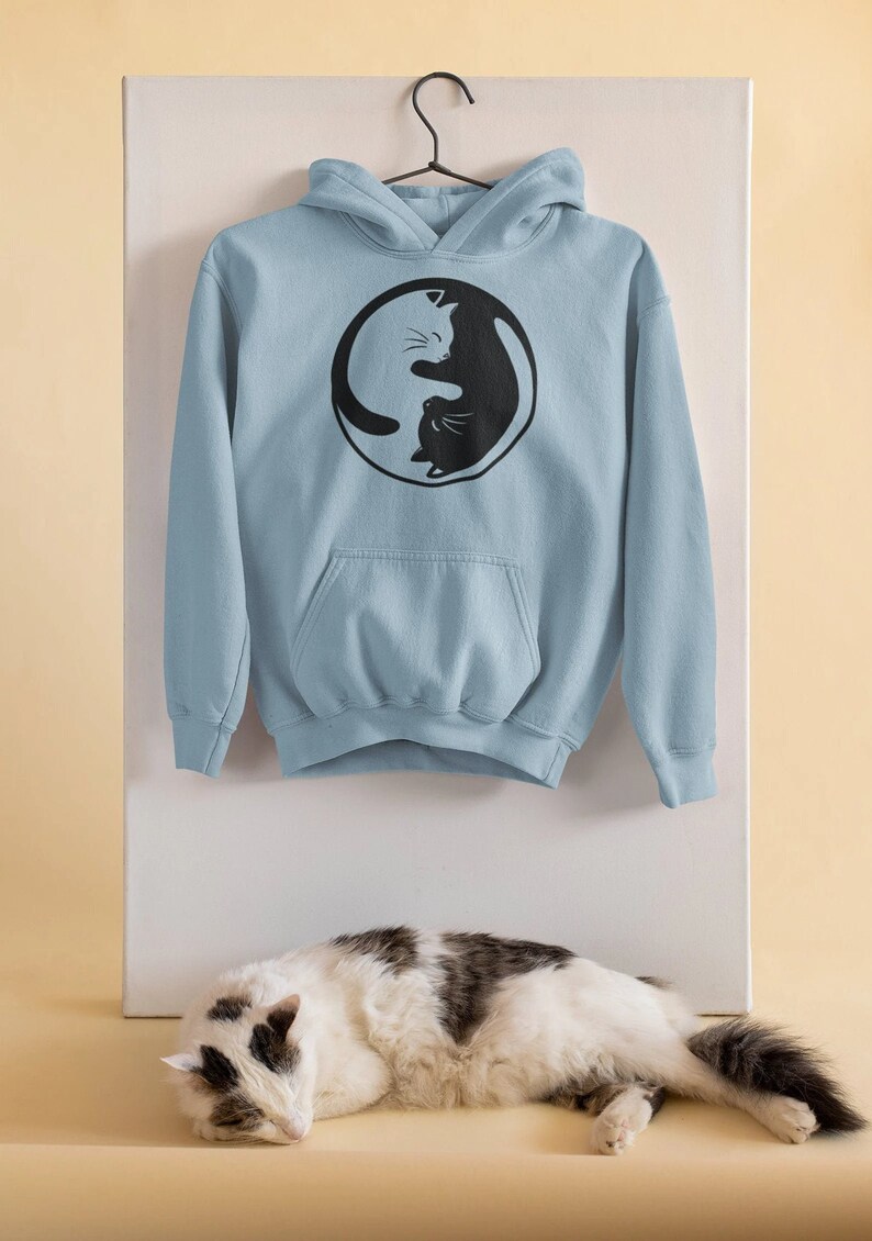 May include: A light blue hoodie with a black and white cat Yin and Yang design. The hoodie has a front pocket and a hood. The design features two cats, one black and one white, in a circular embrace. The hoodie is hanging on a black hanger.