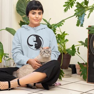 May include: A light blue hoodie with a black and white Yin and Yang cat design. The person is sitting on a tiled floor with a gray cat. The background includes potted plants and a vintage speaker.