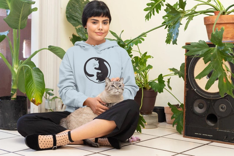 May include: A light blue hoodie with a black and white Yin and Yang cat design. The person is sitting on a tiled floor with a gray cat. The background includes potted plants and a vintage speaker.