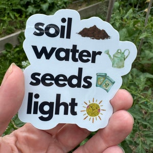 May include: White sticker with black text that reads "soil water seeds light". The sticker has illustrations of dirt, a watering can, seed packets, and a sun.