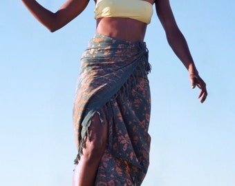 May include: A woman wearing a yellow tube top and a patterned sarong with a fringed edge. She is holding a yellow flower against a clear blue sky. She is also wearing sunglasses and a necklace.