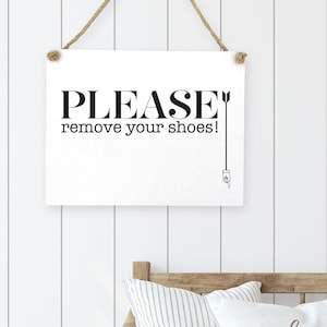 May include: A white rectangular sign with the text "PLEASE remove your shoes!" in black, hanging by a rope. Below, a bench with pillows that say "welcome home" and striped pillows. Green boots are visible below the bench.